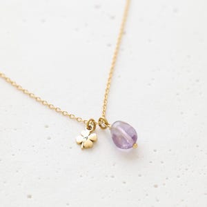 May include: A delicate gold necklace featuring a four-leaf clover charm and a teardrop-shaped amethyst pendant. The chain is fine and the charms are small, creating an elegant and minimalist design. The amethyst has a soft purple hue.