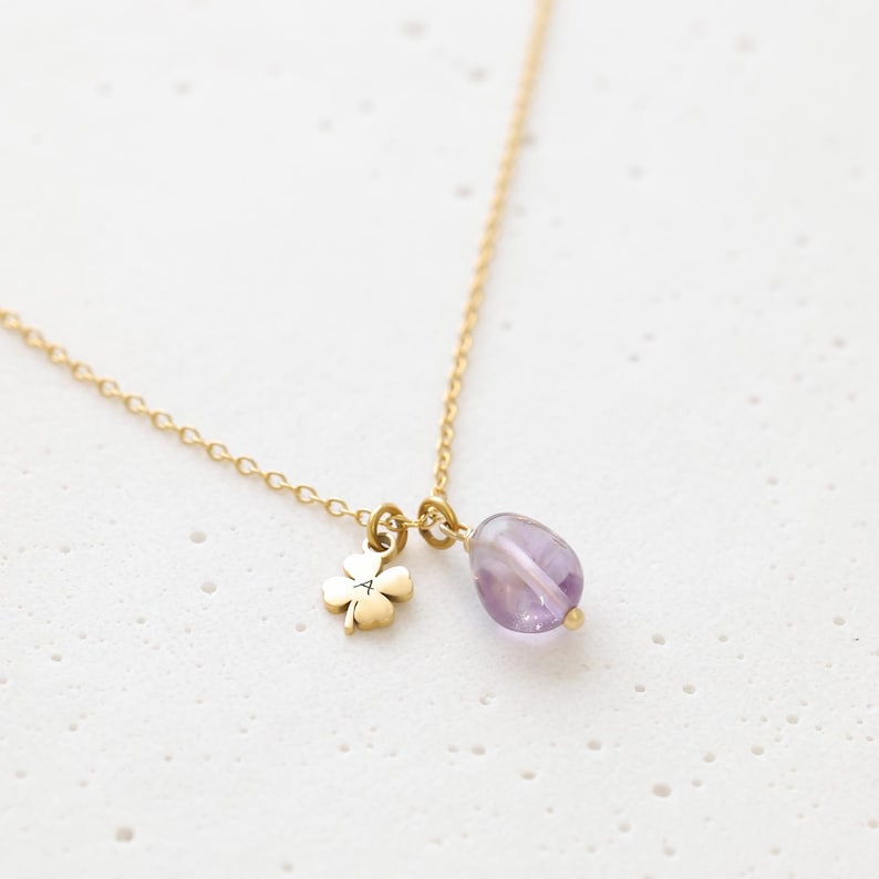 May include: A delicate gold necklace featuring a four-leaf clover charm and a teardrop-shaped amethyst pendant. The chain is fine and the charms are small, creating an elegant and minimalist design. The amethyst has a soft purple hue.