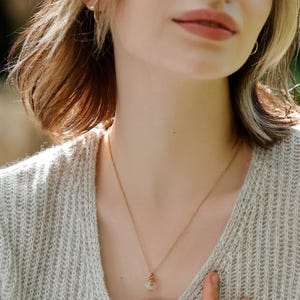 May include: A woman wearing a light beige sweater with a delicate gold chain necklace featuring a small, clear glass pendant.