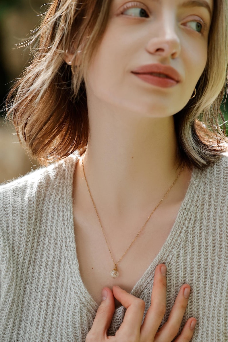 May include: A woman wearing a light beige sweater with a delicate gold chain necklace featuring a small, clear glass pendant.
