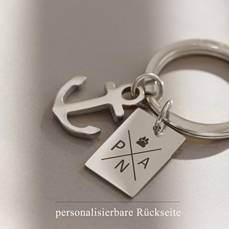 May include: Silver keyring with an anchor charm and a rectangular tag engraved with the letters "PNA" and a paw print. The tag has the text "personalisierbare R&uuml;ckseite" on the back.