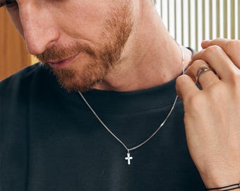 May include: A silver necklace with a small cross pendant. The necklace is worn with a black shirt. A silver ring is on the right hand. A black bracelet is on the right wrist.