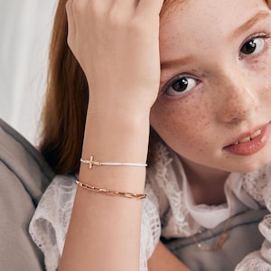 May include: A white string bracelet with a gold cross charm and a gold chain bracelet. The bracelets are worn on a person's wrist.