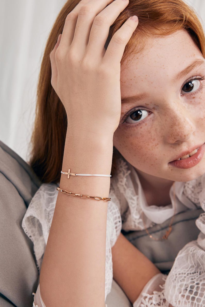 May include: A white string bracelet with a gold cross charm and a gold chain bracelet. The bracelets are worn on a person's wrist.