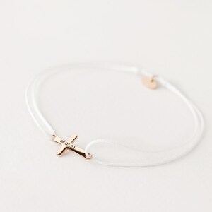 May include: White string bracelet with a gold cross charm engraved with the date "08.05.21".