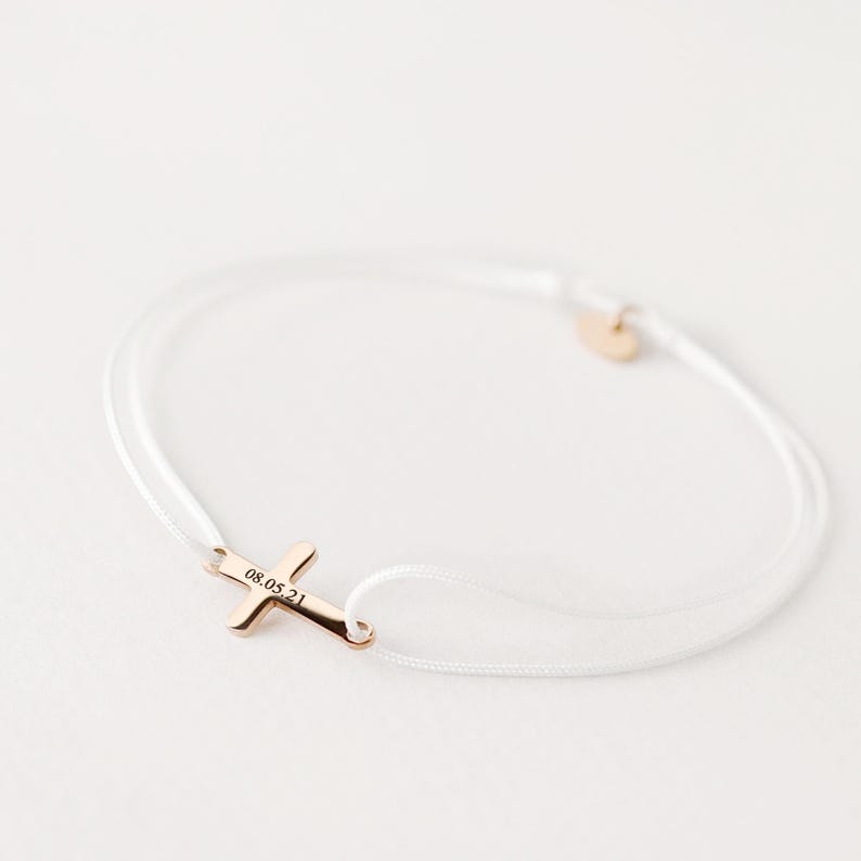 May include: White string bracelet with a gold cross charm engraved with the date "08.05.21".