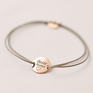 May include: A delicate bracelet with a rose gold charm that reads "Beste Mama" with a small heart symbol. The bracelet is made of a grey cord.