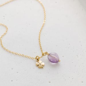 May include: A delicate gold-colored necklace featuring a four-leaf clover charm and a purple amethyst gemstone pendant. The chain is fine and the pendant is a light purple color with white stripes.