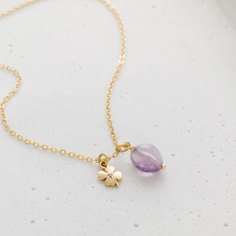 May include: A delicate gold-colored necklace featuring a four-leaf clover charm and a purple amethyst gemstone pendant. The chain is fine and the pendant is a light purple color with white stripes.