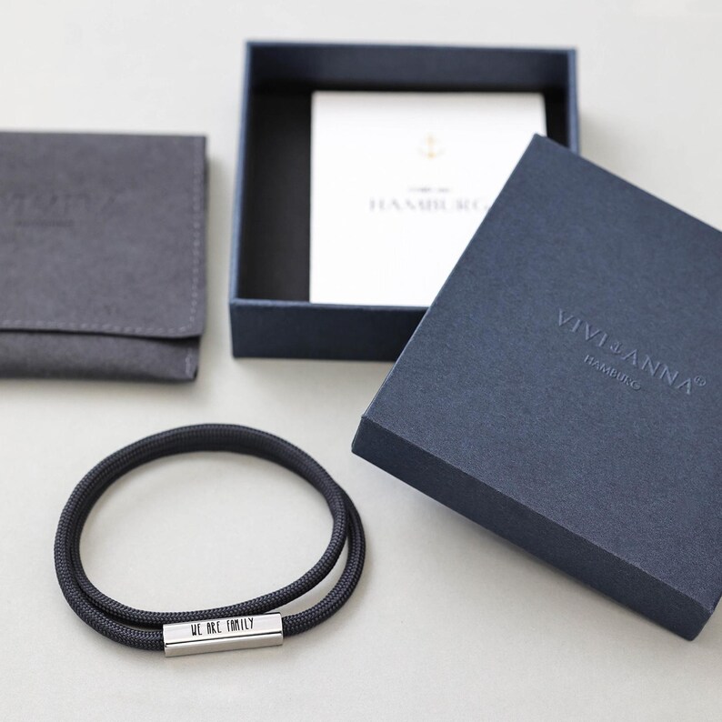 May include: A black cord bracelet with a silver bar that reads "We are family". The bracelet is packaged in a blue box with the Vivianna Hamburg logo.