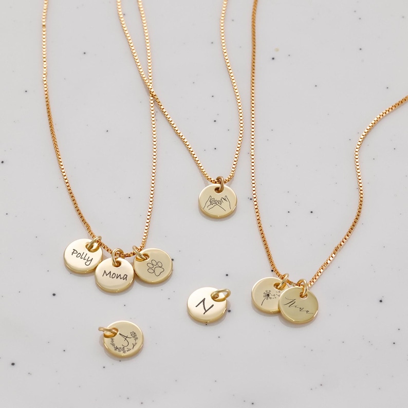 Op de afbeelding: Vier gouden kettingen met verschillende bedels. E&eacute;n ketting heeft een bedel met twee handen die elkaars pink vasthouden, een andere heeft een bedel met een pootprint, een andere heeft drie bedels met de namen "Polly", "Mona" en een pootprint, en de laatste ketting heeft twee bedels met een paardenbloem en de naam "Nora".