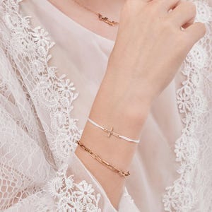 May include: A white string bracelet with a small gold cross charm. The bracelet is worn on a wrist with a gold chain bracelet.
