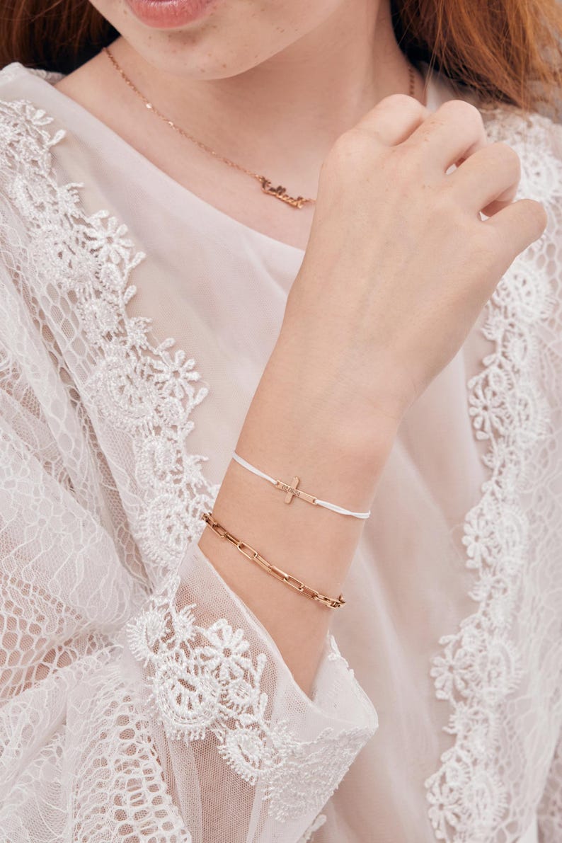 May include: A white string bracelet with a small gold cross charm. The bracelet is worn on a wrist with a gold chain bracelet.