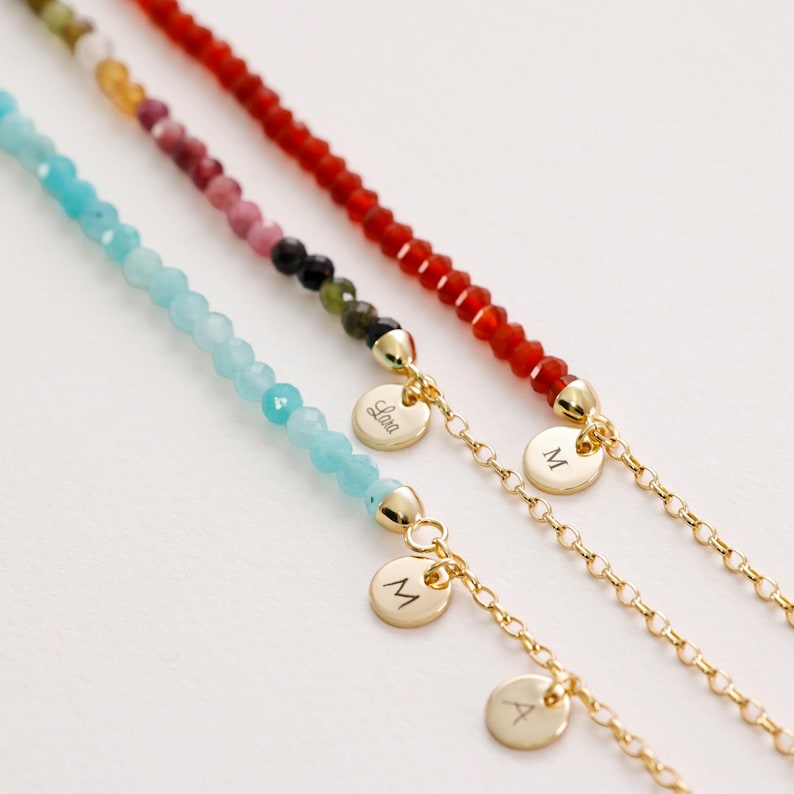 May include: Three gold chain bracelets with personalised charms. The top bracelet has red beads, the middle bracelet has blue beads, and the bottom bracelet has multi-coloured beads. The charms are engraved with the letters "M", "A", and "Lola".