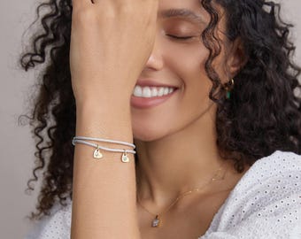 May include: A woman wearing a white shirt with a grey bracelet featuring two gold heart charms. The charms have small engraved letters.