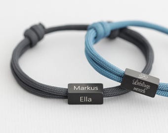 May include: Two adjustable cord bracelets featuring rectangular black metal beads. One dark gray cord bracelet is engraved with the names Markus and Ella. One blue cord bracelet is engraved with the text Lieblingsmensch and a four-leaf clover icon.