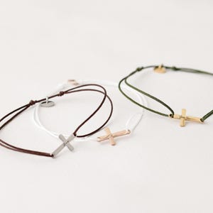 May include: Three bracelets with different colored cords and cross charms. The charms are silver, gold, and rose gold. The cords are brown, white, and green.