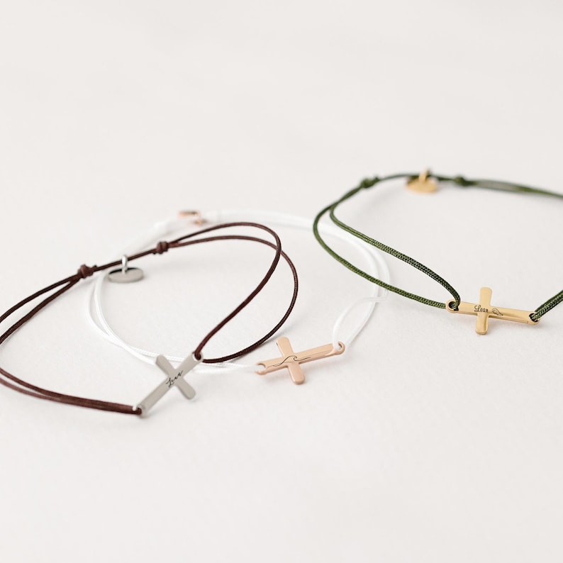 May include: Three bracelets with different colored cords and cross charms. The charms are silver, gold, and rose gold. The cords are brown, white, and green.
