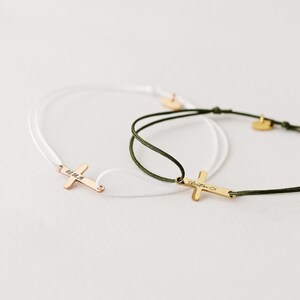 May include: Two simple bracelets with a gold cross charm. One bracelet is white cord and the other is green cord. The cross charm on the white cord bracelet has the inscription "02.04.18" and the cross charm on the green cord bracelet has the inscription "Christina".