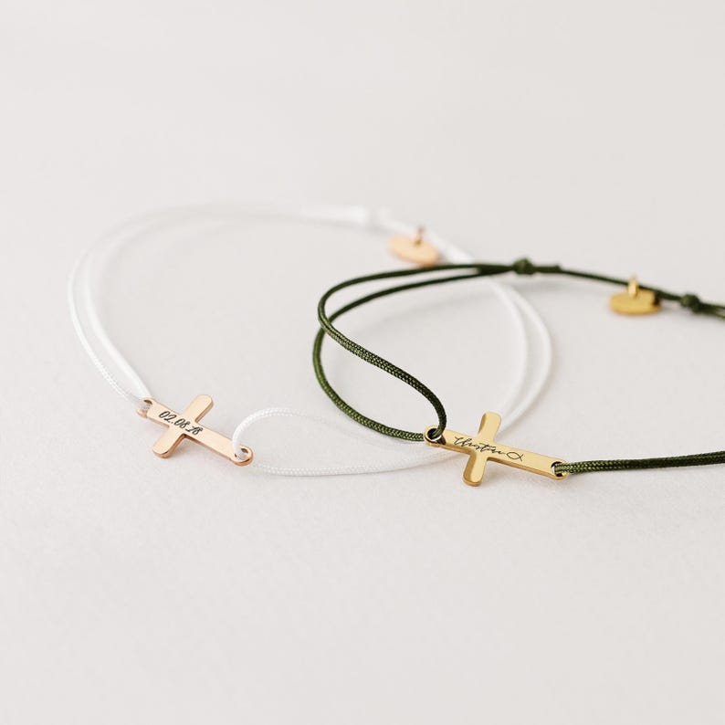 May include: Two simple bracelets with a gold cross charm. One bracelet is white cord and the other is green cord. The cross charm on the white cord bracelet has the inscription "02.04.18" and the cross charm on the green cord bracelet has the inscription "Christina".