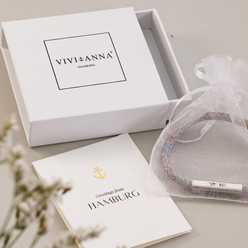 May include: A white box with the text "VIVI+ANNA HAMBURG" and a greeting card that says "Greetings from HAMBURG" with a gold anchor. A colorful bracelet is in a white mesh bag.