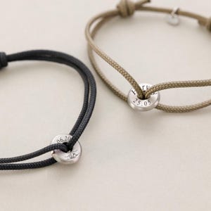 May include: Two friendship bracelets with black and beige cords and silver metal beads. The beads are engraved with the dates "06.20" and "25.05".