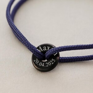 May include: A dark blue braided cord bracelet with a black metal tag engraved with the name "Max" and the date "20.04.2025".