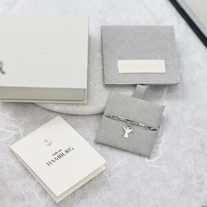 May include: A white box with the text "Liebe aus HAMBURG" on a white card. The box is open and a small grey pouch with a silver chain and angel charm is inside. The pouch is on a white surface.