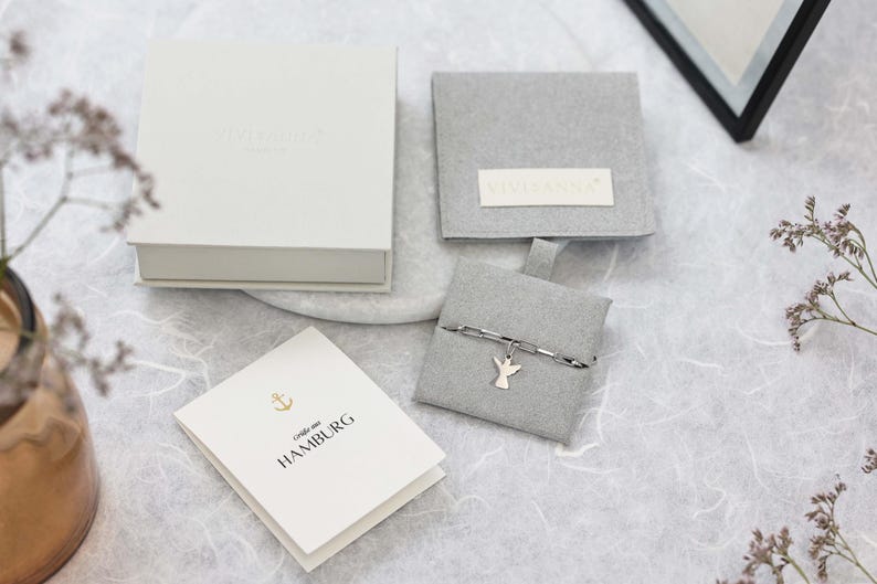 May include: A white box with the text "Liebe aus HAMBURG" on a white card. The box is open and a small grey pouch with a silver chain and angel charm is inside. The pouch is on a white surface.