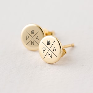 Può includere: Un paio di orecchini a bottone color oro. Ogni orecchino è rotondo con un'impronta di zampa e le lettere P, A e N disposte a croce. Gli orecchini sono su una superficie bianca.