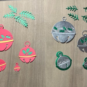 May include: Assortment of red and silver jingle bells with green holly leaf accents. The bells are in various sizes and are arranged on a wooden surface.
