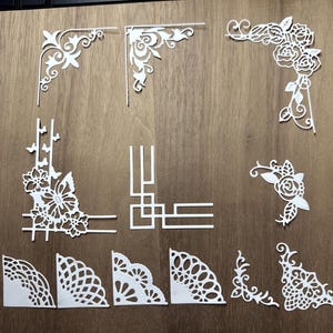 May include: A collection of white paper corner embellishments in various floral and geometric designs. The assortment includes delicate roses, butterflies, and abstract patterns, perfect for scrapbooking or card making. The corners are arranged on a wooden surface.