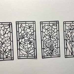 May include: Four black paper cutouts of stained glass windows with floral designs. Each window features a different flower design, including lilies, roses, and irises.