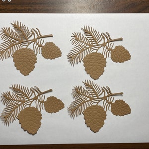 May include: Four brown paper pine cone and branch cutouts on a white background. The cutouts are intricately detailed and have a rustic, natural look.