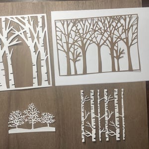 May include: Four white paper cutouts of birch trees, each with a different design. The cutouts are on a brown wooden surface.