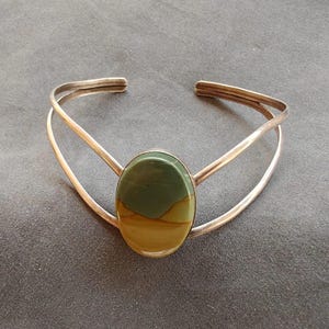 May include: Silver cuff bracelet featuring an oval-shaped stone with green and tan hues. The stone is set in a silver bezel, and the bracelet has a simple, open design. The bracelet is a piece of jewelry.