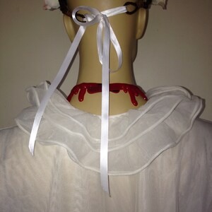 May include: A white, ruffled collar with a white ribbon tied in a bow at the back of the neck. The ribbon is attached to a white flower crown with red fake blood dripping down the back of the neck.