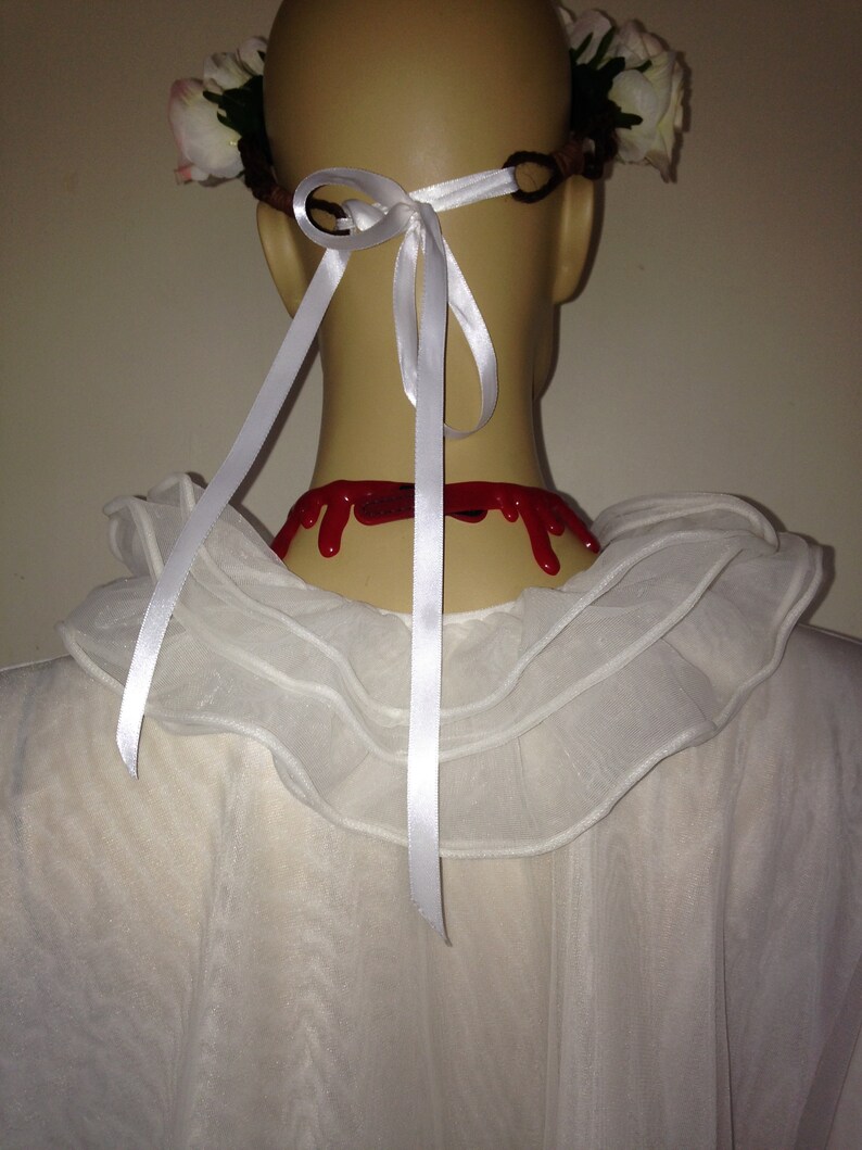 May include: A white, ruffled collar with a white ribbon tied in a bow at the back of the neck. The ribbon is attached to a white flower crown with red fake blood dripping down the back of the neck.