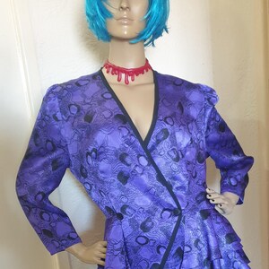 May include: A purple and black patterned blouse with a ruffled hem and a V-neckline. The blouse has a black trim and a black sash. The model is wearing a red dripping necklace.