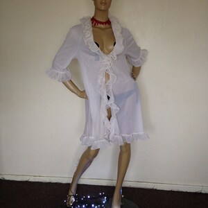 May include: A white sheer robe with ruffled trim and a tie at the waist. The robe is worn by a mannequin with a flower crown and a red choker necklace. The mannequin is standing on a black surface with white string lights.
