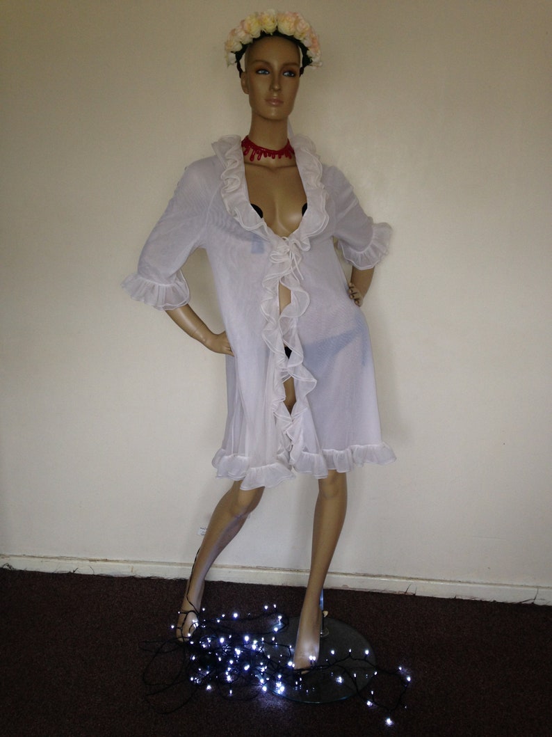 May include: A white sheer robe with ruffled trim and a tie at the waist. The robe is worn by a mannequin with a flower crown and a red choker necklace. The mannequin is standing on a black surface with white string lights.