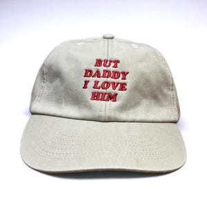 May include: A beige baseball cap with red embroidered text that reads "But Daddy I Love Him".