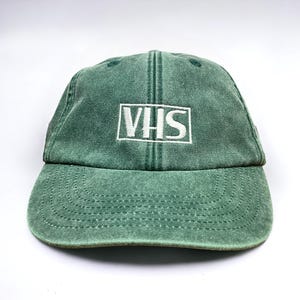 May include: A green baseball cap with a white embroidered logo that says "VHS".