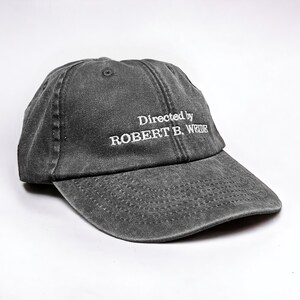 May include: A black, distressed, cotton baseball cap with white embroidered text that reads "Directed by ROBERT B. WEIDE".