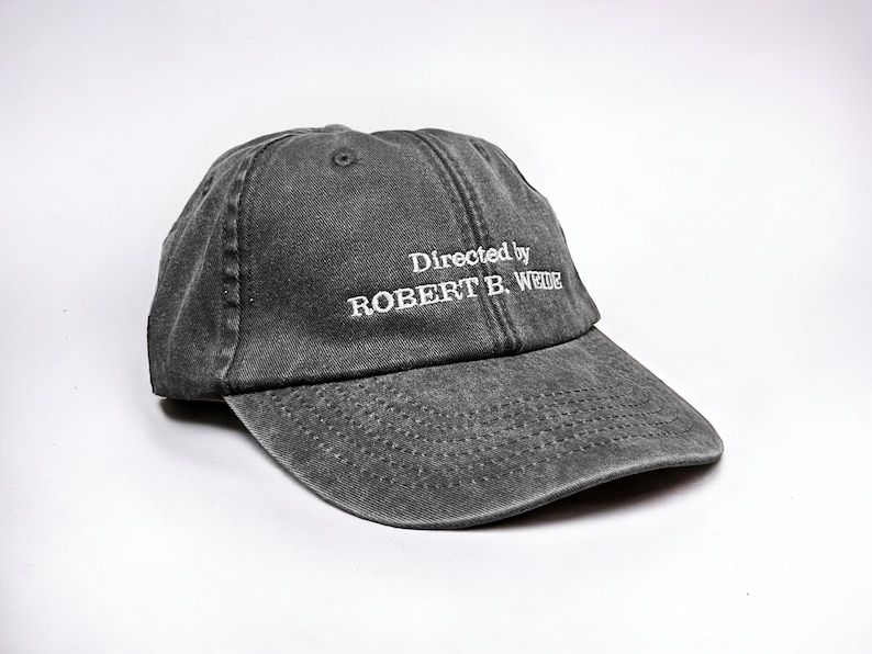 May include: A black, distressed, cotton baseball cap with white embroidered text that reads "Directed by ROBERT B. WEIDE".