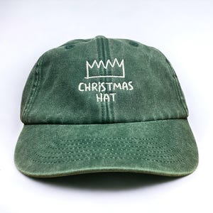 以下が含まれることがあります： 白い刺繍で「CHRISTMAS HAT」の文字と白い王冠のデザインが施された、グリーンの野球帽。 つばがカーブしており、質感のある生地で作られています。