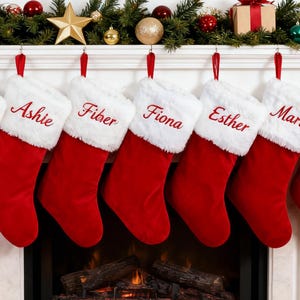 May include: Seven red velvet Christmas stockings with white faux fur cuffs hanging from a fireplace mantel. Each stocking is embroidered with a name in red script: Steven, Ashie, Fiber, Fiona, Esther, Maria, and Charlie. Green garland and ornaments decorate the mantel above.