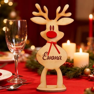 Puede incluir: Decoración de mesa navideña de reno de madera con el nombre "Emma" grabado en el cuerpo. El reno tiene una nariz roja y una cinta roja. El fondo incluye velas, una copa de vino y un mantel rojo.