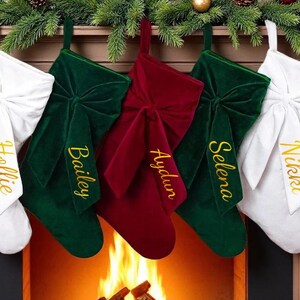 May include: Five Christmas stockings hang above a fireplace. Two white stockings with bows flank green and red velvet stockings. Each stocking has a name embroidered in gold. A garland with pinecones and red baubles decorates the mantelpiece.