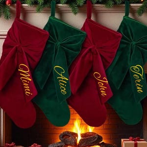 May include: Four Christmas stockings, two red and two green, hang above a fireplace with a burning fire. Each stocking has a bow and a name embroidered in gold. The scene is decorated with garland and wrapped gifts.